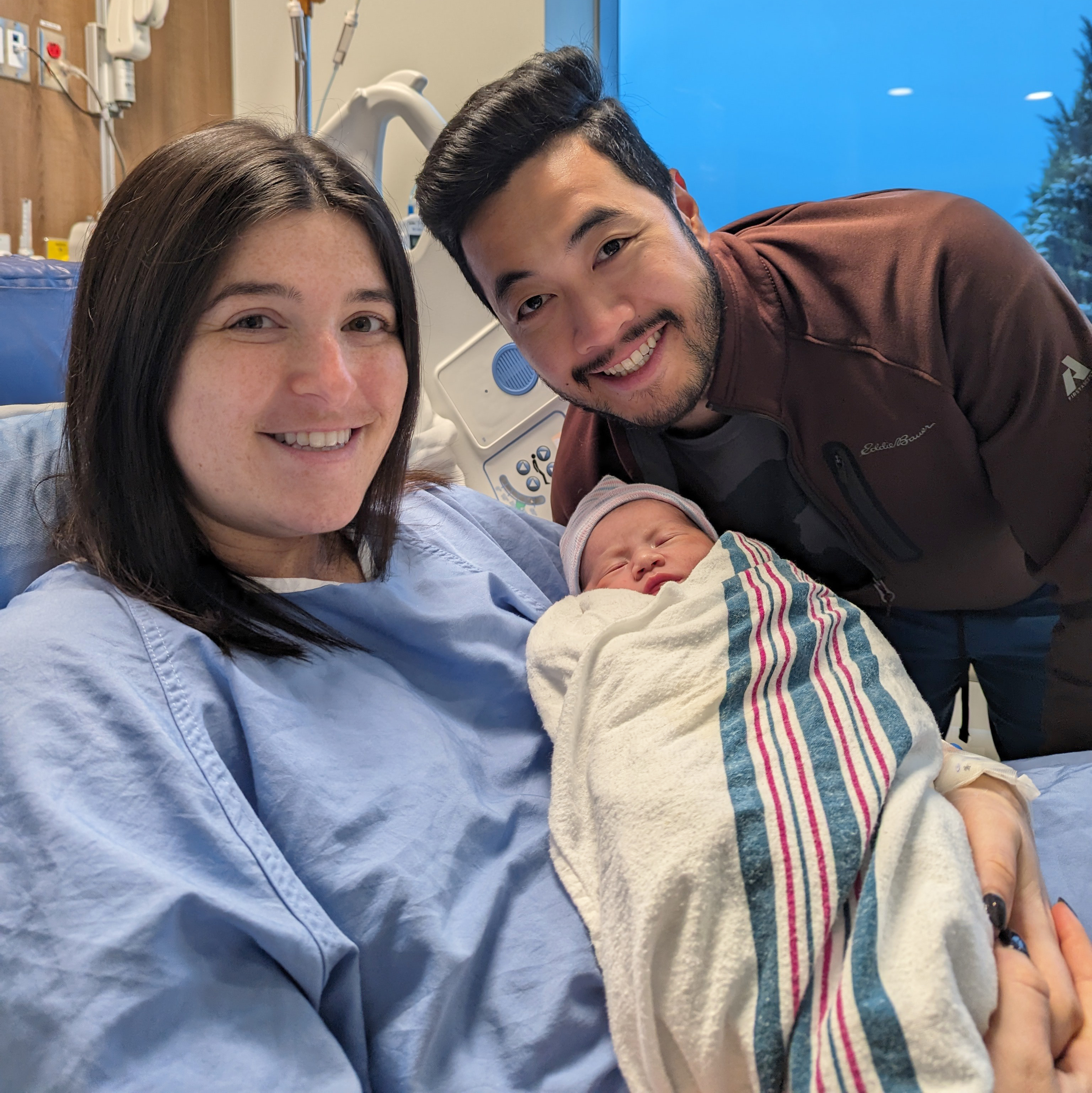 An image of new parents, Vic and Katie with their new baby. Katie is in the hospital bed with Vic standing beside her. Katie is holding the baby.