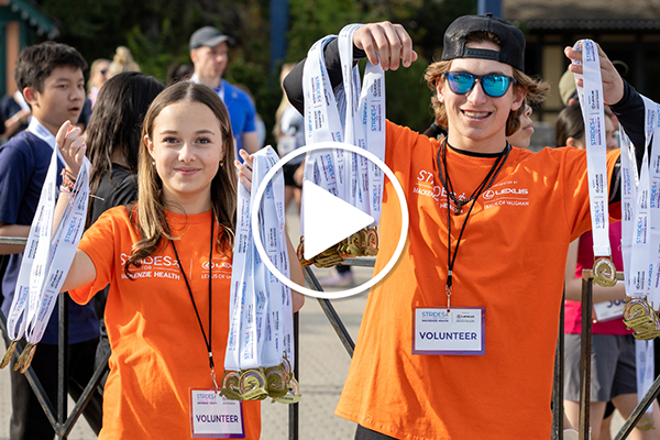 Image of two volunteers handing out medals at Strides for Mackenzie Health. A play symbol is present indicating an embedded link to our volunteer week video.