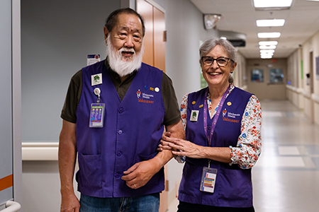 Two smiling volunteers in purple vests in a hospital hallway